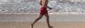 Lifeguard running shirtless on the beach in red shorts close to the water