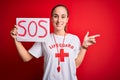 Lifeguard woman wearing t-shirt with red cross and whistle holding banner with sos message very happy pointing with hand and