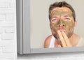 Lifestyle bathroom mirror portrait of young attractive happy man with green cream on his face applying facial mask sending kiss to Royalty Free Stock Photo