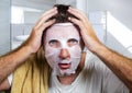 Young scared and surprised man at home using beauty paper facial mask cleansing doing anti aging facial treatment looking himself Royalty Free Stock Photo
