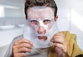 Young scared and surprised man at home using beauty paper facial mask cleansing doing anti aging facial treatment looking himself Royalty Free Stock Photo
