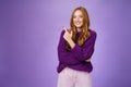 Charming friendly-looking bright redhead woman with freckles and makeup in purple warm sweater pointing at upper left Royalty Free Stock Photo