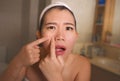 Lifestyle face portrait of young upset and expressive Asian Chinese woman squeezing pimples while looking at the mirror in the Royalty Free Stock Photo