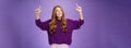 Dreamy girl with red hair and freckles in warm cozy purple sweater raising hands looking and pointing up with intrigued