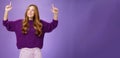 Dreamy girl with red hair and freckles in warm cozy purple sweater raising hands looking and pointing up with intrigued