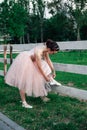lifestyle full-length portrait of a ballerina in a pink dress with a full organza skirt bends down to her leg to tie