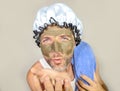 Lifestyle funny portrait of happy weird man on shower cap kissing to himself in bathroom mirror with green cream on his face apply