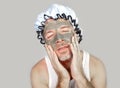 Lifestyle funny portrait of happy weird man on shower cap looking to himself in bathroom mirror with green cream on his face apply