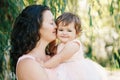 Lifestyle group portrait of beautiful white Caucasian brunette mother holding hugging daughter in pink dress kissing her in cheek