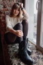 Lifestyle natural light portrait young adorable woman in white cozy sweater black boots tights sits on a windowsill and looks at