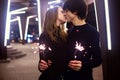 Lifestyle portrait of couple in love holding sparkling New Year fireworks on the city streets with lot of lights on background. Royalty Free Stock Photo