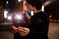 Lifestyle portrait of couple in love holding sparkling New Year fireworks on the city streets with lot of lights on background. Royalty Free Stock Photo