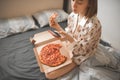 Lifestyle portrait of a girl in the bedroom, sitting on a bed with a box of pizza, using the internet on a smartphone, eating a Royalty Free Stock Photo