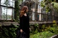 Lifestyle portrait of girl wearing blank black t-shirt, jeans and coat posing against building covered with green leaves Royalty Free Stock Photo