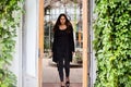 Lifestyle portrait of girl wearing blank black t-shirt, jeans and coat posing against building covered with green leaves Royalty Free Stock Photo