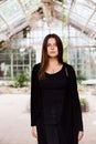 Lifestyle portrait of girl wearing blank black t-shirt, jeans and coat posing against building covered with green leaves Royalty Free Stock Photo