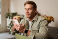 Lifestyle portrait of handsome young man, sitting in cafe, checking his phone and drinking coffee, smiling. Royalty Free Stock Photo