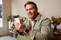 Lifestyle portrait of handsome young man, sitting in cafe, checking his phone and drinking coffee, smiling. Royalty Free Stock Photo