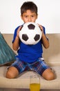 Lifestyle portrait at home of young 7 or 8 years old boy holding soccer ball watching excited and nervous football game on televis