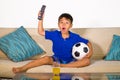 Lifestyle portrait at home of young 7 or 8 years old boy holding soccer ball watching excited and nervous football game on televis