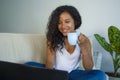 Young attractive and relaxed hispanic student woman sitting at home sofa couch networking with laptop computer drinking coffee or Royalty Free Stock Photo