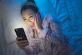 Lifestyle portrait of young beautiful happy and relaxed Asian Korean woman on bed laughing with mobile phone on bed in the dark in Royalty Free Stock Photo