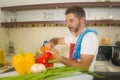 Lifestyle portrait of young handsome and happy man in red apron cutting carrot preparing vegetables salad smiling cheerful in home Royalty Free Stock Photo