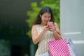 Young happy and beautiful Asian Chinese woman walking on the street carrying shopping bags using mobile phone searching online for Royalty Free Stock Photo