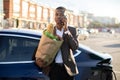 Smiling young afro american businessman, holding paper bag with food, talking on mobile phone and looking at camera Royalty Free Stock Photo