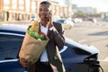 Smiling young afro american businessman, holding paper bag with food, talking on mobile phone and looking at camera Royalty Free Stock Photo