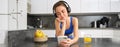 Lifestyle and workout. Young smiling woman in headphones, standing in kitchen with smartphone, drinking orange juice and Royalty Free Stock Photo