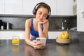 Lifestyle and workout. Young smiling woman in headphones, standing in kitchen with smartphone, drinking orange juice and Royalty Free Stock Photo