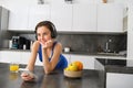 Lifestyle and workout. Young smiling woman in headphones, standing in kitchen with smartphone, drinking orange juice and Royalty Free Stock Photo