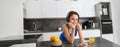 Lifestyle and workout. Young smiling woman in headphones, standing in kitchen with smartphone, drinking orange juice and Royalty Free Stock Photo