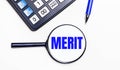 On a light background, a black calculator, a blue pen and a magnifying glass with text inside the MERIT. View from above Royalty Free Stock Photo