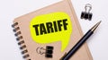 On a light background, a brown notebook, a black pen and paper clips, and a yellow card with the text TARIFF. Business concept