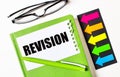 On a light background, multi-colored arrows, black glasses, a green notebook with a pen and a white card with the text REVISION