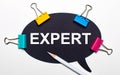 On a light background, multi-colored paper clips, a white pencil and black paper with the words EXPERT. View from above Royalty Free Stock Photo