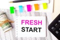 On a light background, a pen, dollars, a notebook with the text FRESH START and bright stickers. Business concept