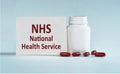 On a light blue background a card with the text NHS - NATIONAL HEALTH SERVICE near the white bottle pills Royalty Free Stock Photo