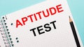 On a light blue background, a checkered notebook with the words APTITUDE TEST and a black pencil Royalty Free Stock Photo