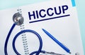 On a light blue background, an open notebook with the word HICCUP, a blue pen and a stethoscope. View from above. Medical concept