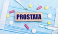 On a light blue disposable face mask there are tablets, a thermometer, an ampoule and a wooden block with the text PROSTATA.