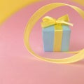 Light blue gift box at the end of the spiral yellow ribbon, pink background, square. Royalty Free Stock Photo