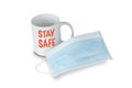 A light blue medical face mask in front of a white mug with red text saying STAY SAFE on white background