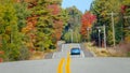 Light blue Mustang cruises down the empty road running through colorful forest.