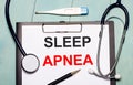 On a light blue wooden background, there is a paper labeled SLEEP APNEA, a stethoscope, an electronic thermometer, and a pen.