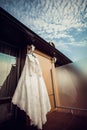 A light-colored wedding dress hangs by the hotel window against a blue sky with clouds Royalty Free Stock Photo