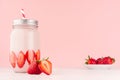 Light dairy strawberry drink in trendy jar with ripe pieces berry on saucer, red striped straw, silver lid on gentle pink color.