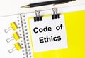 On a light desktop, a pen, yellow paper clips and a white card with the text CODE OF ETHICS on a yellow background